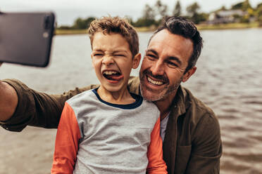 Nahaufnahme von Vater und Sohn, die in der Nähe eines Sees ein Selfie machen. Ein glückliches Kind macht Grimassen und streckt die Zunge heraus, während sein Vater ein Selfie macht. - JLPSF22946