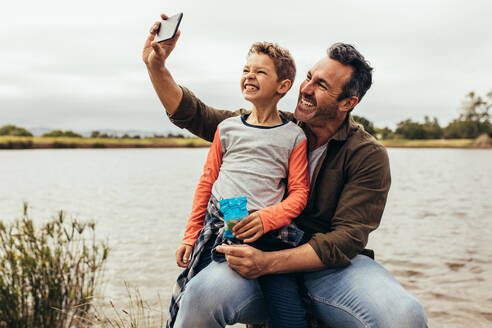 Ein Mann macht ein Selfie mit seinem Sohn an einem See. Ein lächelnder Vater und sein Sohn machen ein Selfie bei einem Ausflug zu einem See. - JLPSF22947