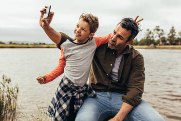 Mann sitzt mit seinem Sohn auf einem Baumstamm an einem See und macht ein Selfie. Mann genießt seine Freizeit, während er mit seinem Sohn an einem See sitzt. - JLPSF22948