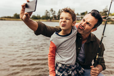 Close up of father and son taking a selfie while fishing near a lake. Happy father and son making faces while takes a selfie near the lake holding a fishing rod. - JLPSF22949