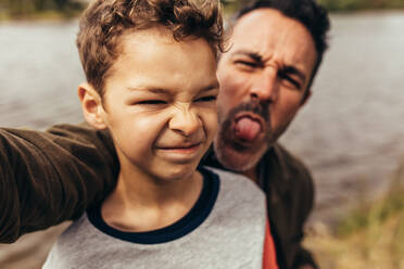 Close up of a kid standing near a lake with his father making faces standing behind him. Father and son spending time together having fun outdoors. - JLPSF22950