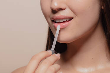 Close up of young korean woman applying transparent lip gloss. Cropped shot of girl putting on makeup on her lips with applicator against beige background. - JLPSF22952