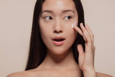 Nahaufnahme eines asiatischen weiblichen Modells, das Feuchtigkeitscreme auf ihr Gesicht aufträgt und wegschaut. Frau, die Feuchtigkeitscreme auf ihr hübsches Gesicht vor beigem Hintergrund aufträgt. - JLPSF22961
