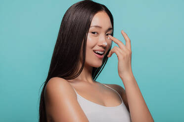 Asian female model applying moisturizer to her nose and looking at camera. Woman applying moisturizer cream on her face against blue background. - JLPSF22974