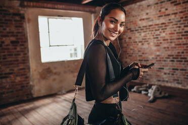 Portrait of a young woman in black sportswear listening to music from smart phone in hands and carrying a sports bag. Fitness woman at gym looking at camera and smiling. - JLPSF22997