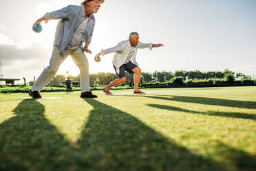 Menschen spielen auf einer Wiese mit Sonne im Hintergrund Boule. Zwei ältere Personen beugen sich vor, um Boule zu werfen, wobei ihr Schatten auf dem Boden liegt. - JLPSF23008