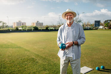 Smiling old man standing in a park with a boules in hand. Cheerful elderly man in hat standing in a play ground holding a boule. - JLPSF23016