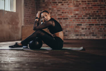 Junge Frau sitzt mit einem Medizinball auf dem Boden eines Fitnessstudios und ruht sich nach dem Training im Fitnessstudio aus. - JLPSF23017