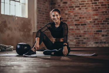 Porträt einer jungen Frau während einer Pause im Fitnessstudio. Hispanische Frau sitzt auf dem Boden und schaut in die Kamera, ruht sich nach dem Training aus. - JLPSF23018