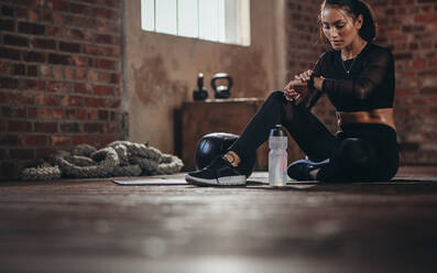 Eine fitte junge Frau sitzt auf dem Boden und schaut auf ihre Smartwatch im Fitnessstudio. Eine Sportlerin schaut sich nach dem Training ihre Leistung auf der Smartwatch-Fitness-App an. - JLPSF23022