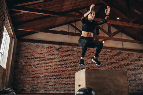 Aufnahme einer jungen Frau, die im Rahmen eines Trainings auf eine Box springt. Fitness-Frau beim Box-Jump-Workout im Fitnessstudio. - JLPSF23034
