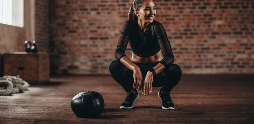 Lächelnde Frau hockt im Fitnessstudio mit einem Medizinball auf dem Boden. Fitness-Frau macht Pause nach dem Training im Fitnessstudio. - JLPSF23037