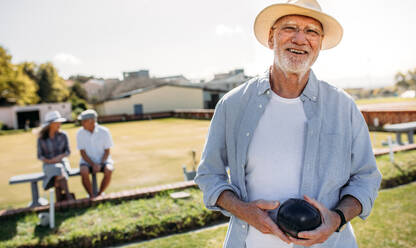 Happy senior man standing in a lawn holding a boules. Elderly man in hat playing boules in a park while his friends are sitting in the background. - JLPSF23085