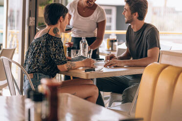 Junges Paar sitzt in einem Café und gibt einer Kellnerin eine Bestellung auf. Ein Mann gibt einer Kellnerin im Restaurant eine Bestellung auf, während er mit seiner Freundin zusammensitzt. - JLPSF23089