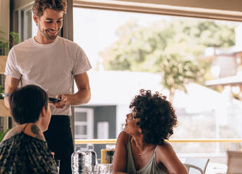 Junge Frau mit einer Freundin bestellt bei einem Kellner im Café. Zwei Frauen sitzen im Café und geben dem männlichen Kellner eine Bestellung auf. - JLPSF23090