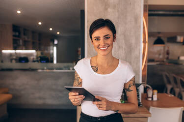 Portrait of a smiling coffee shop owner standing inside her shop. Female restaurant owner standing with a digital tablet. - JLPSF23094