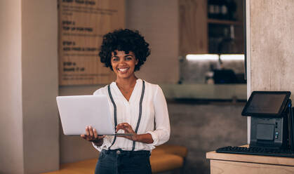 Porträt einer jungen afrikanischen Frau mit einem Laptop in ihrem Restaurant. Cafébesitzerin mit Laptop und lächelnd. - JLPSF23102