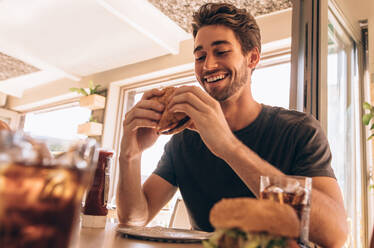 Glücklicher junger Mann, der einen leckeren Burger im Restaurant isst. Hungriger junger Mann, der einen gestapelten Burger in einem Fast-Food-Café isst. - JLPSF23130