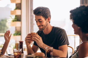 Young man eating burger while sitting with female friends at a restaurant. Man hanging out at cafe with friends and eating burger. - JLPSF23132