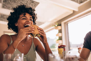 African woman eating stack burger at restaurant with friends. Happy young woman having junk food at cafe with friends. - JLPSF23134