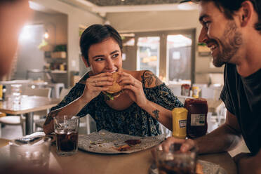 Junge Frau isst Burger, während sie mit Freunden im Restaurant sitzt. Junge Leute unterhalten sich und essen Burger im Café. - JLPSF23137
