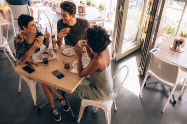 Blick von oben auf drei junge Freunde, die in einem Restaurant einen Burger essen. Kleine Gruppe von Männern und Frauen, die um einen Tisch im Café sitzen, sich unterhalten und essen. - JLPSF23139
