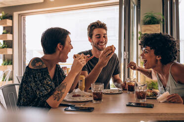 Eine Gruppe von Freunden isst einen Burger in einem Restaurant. Fröhliche junge Leute essen einen Burger und genießen ihn in einem Fast Food Restaurant. - JLPSF23140