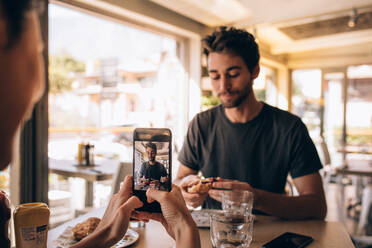 Woman sitting at a restaurant taking photograph of her boyfriend eating a burger. Woman shooting pictures with mobile phone of man eating burger at a restaurant. - JLPSF23141