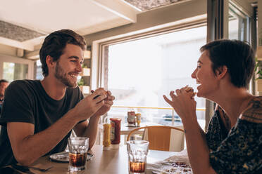 Glückliches Paar isst Burger und genießt im Café. Junger Mann spricht mit seiner Freundin und lächelt beim Essen in einem Restaurant. - JLPSF23144