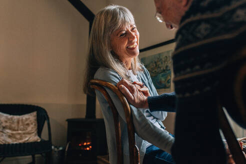 Lächelnde ältere Frau sitzt auf einem Stuhl zu Hause mit einem Mann, der ihren Arm hält. Älteres Paar verbringt Zeit miteinander und spricht zu Hause. - JLPSF23150