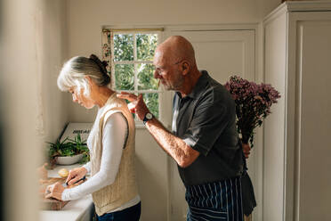 Seitenansicht einer Frau bei der Arbeit in der Küche, hinter der ihr Mann mit einem Blumenstrauß steht. Ein älterer Mann versteckt einen Blumenstrauß hinter sich, um seine Frau zu überraschen. - JLPSF23151