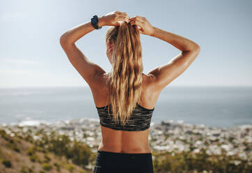 Rückansicht einer fitten jungen Frau, die auf einem Bergpfad steht und sich die Haare zusammenbindet. Läuferin in Sportkleidung auf einer Langlaufloipe, die nach einem Lauf eine Pause macht. - JLPSF23160