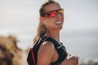 Porträt einer glücklichen jungen Bergläuferin, die in die Kamera lächelt. Attraktive Frau in Sportkleidung und Sonnenbrille. - JLPSF23169