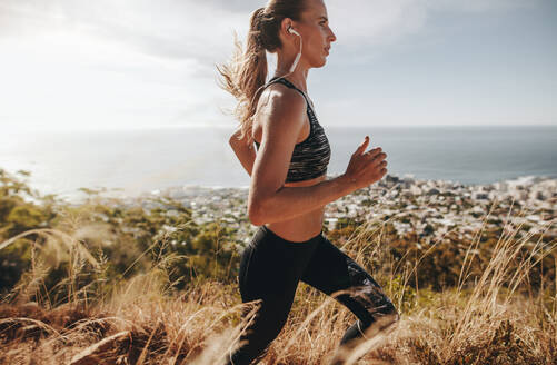 Side view of fit young woman wearing earphones running through mountain trail. Female runner training over rough trail in hillside. - JLPSF23173