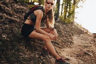 Frau in Sportkleidung sitzt auf einem Felsen und ruht sich nach dem Lauftraining auf einem Bergpfad aus. Trailrunnerin macht Pause vom Training. - JLPSF23185