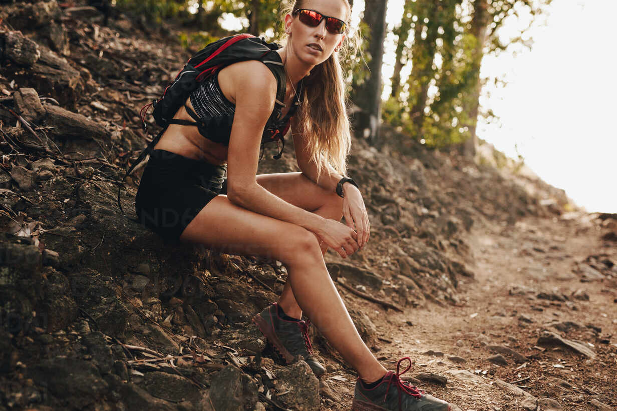 Woman in sportswear sitting on the rock and resting after running training  on mountain trail. Trail runner taking break from training. stock photo