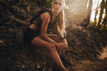 Young woman in sportswear sitting by mountain trail and looking away. Female runner taking a break after running workout. - JLPSF23190