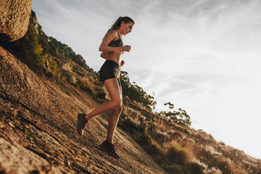 Sporty woman running downhill on a rocky mountain trail. Woman trail runner training for down hill run. - JLPSF23201