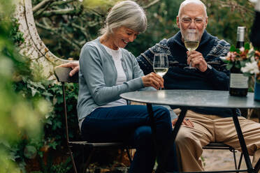 Happy senior couple drinking wine sitting at a table in their backyard. Old woman sitting with her husband drinking wine and talking. - JLPSF23204