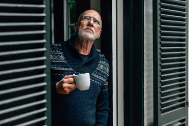 Alter Mann, der mit einer Kaffeetasse in der Hand am Fenster steht und nach oben schaut. Älterer Mann, der zu Hause am Fenster stehend eine Tasse Kaffee genießt. - JLPSF23207