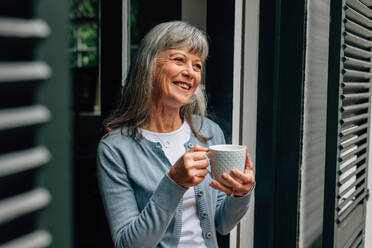 Ältere Frau, die in der Nähe der Tür steht und Kaffee trinkt. Lächelnde alte Frau, die Kaffee trinkt und aus der Tür schaut. - JLPSF23209