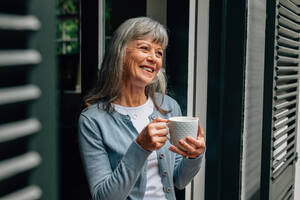 Ältere Frau, die in der Nähe der Tür steht und Kaffee trinkt. Lächelnde alte Frau, die Kaffee trinkt und aus der Tür schaut. - JLPSF23209