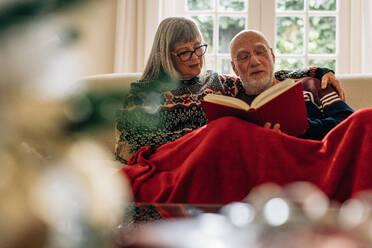 Älteres Ehepaar, das sich beim Lesen eines Buches zu Hause entspannt. Alter Mann und Frau sitzen zusammen auf einer Couch, die mit einer Decke bedeckt ist. - JLPSF23218