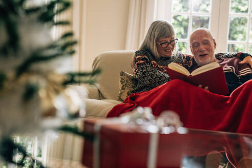 Senior couple sitting on a sofa enjoying reading a book with a gift box in the foreground. Smiling couple spending time together reading a book covering themselves in a blanket at home. - JLPSF23219