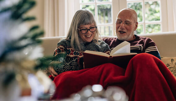 Glücklicher älterer Mann und Frau, die ein Buch lesen und lächeln. Älteres Paar, das Zeit miteinander verbringt, indem es ein Buch auf der Couch zu Hause liest. - JLPSF23220