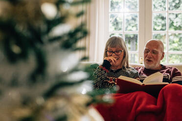 Old woman laughing while her husband reads a book. Senior couple reading a book sitting on couch at home with a christmas tree in the foreground. - JLPSF23221