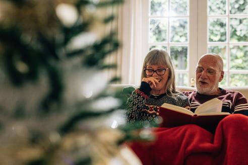 Alte Frau lacht, während ihr Mann ein Buch liest. Älteres Ehepaar liest ein Buch auf der Couch zu Hause mit einem Weihnachtsbaum im Vordergrund. - JLPSF23221