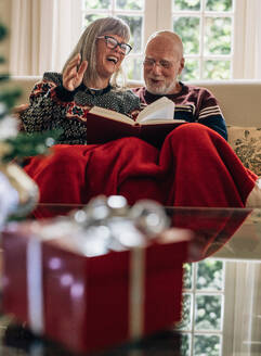 Glückliches älteres Paar, das zu Hause sitzt und ein Buch liest, mit einem Weihnachtsgeschenk im Vordergrund. Älteres Paar, das zu Hause sitzt und ein Buch liest. - JLPSF23222