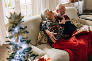 Älteres Paar verbringt glückliche Zeit zu Hause und liest ein Buch zusammen mit einem geschmückten Weihnachtsbaum im Vordergrund. Ältere Frau sitzt mit ihrem Mann und lacht beim Lesen eines Buches. - JLPSF23223