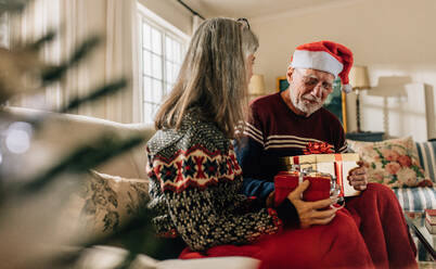 Old couple celebrating christmas at home and exchanging presents. Senior man in santa cap sitting on couch at home with wife holding christmas gifts. - JLPSF23226